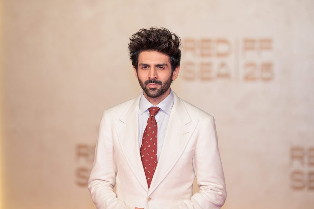 Kartik Aaryan in a white suit and red tie posing on the red carpet
