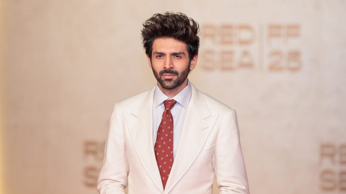 Kartik Aaryan in a white suit and red tie posing on the red carpet