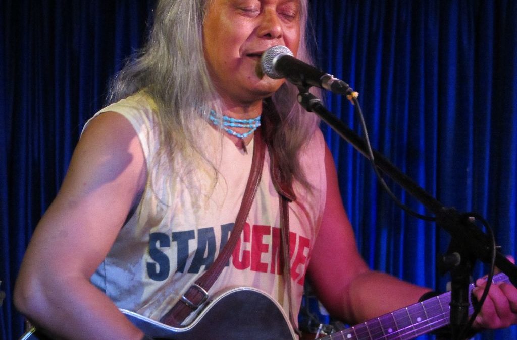 Lou Majaw with long white hair playing a guitar on stage