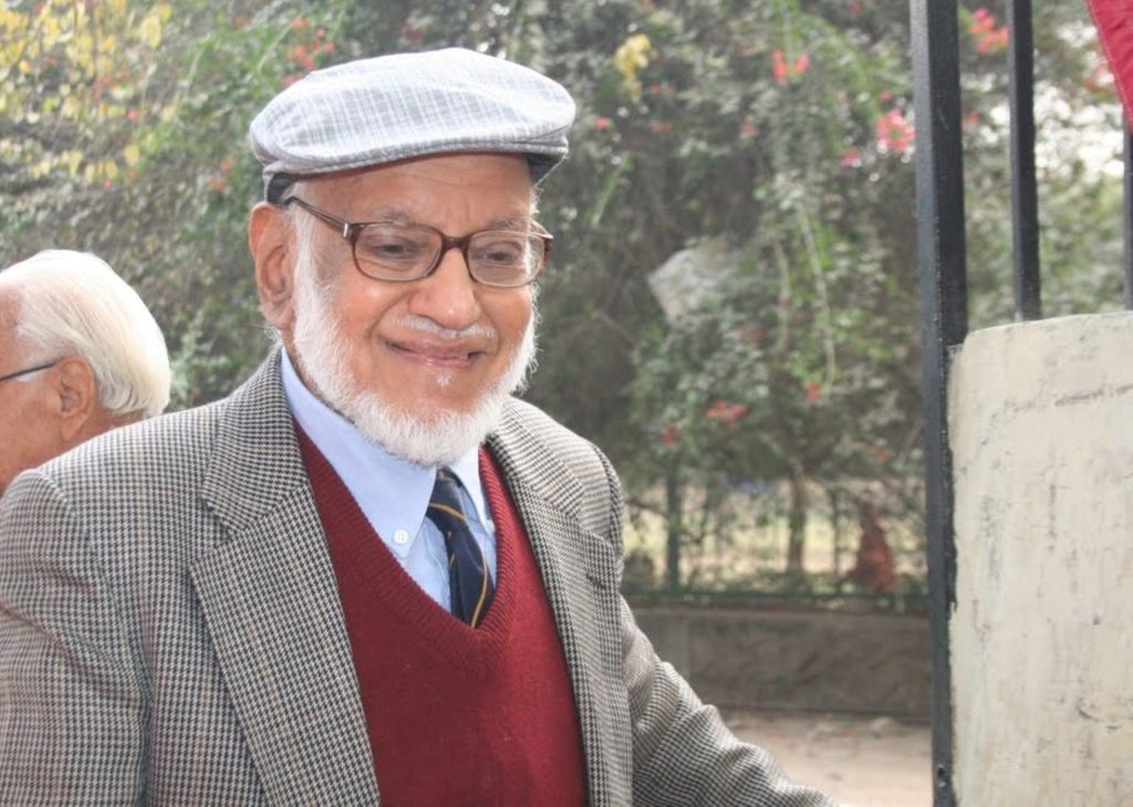 An elderly M. G. K. Menon with a white beard and glasses, wearing a cap and a tweed jacket
