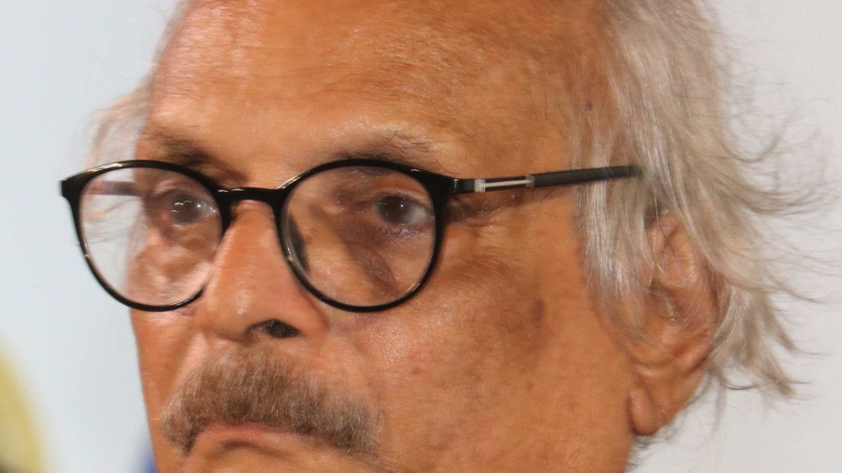M. Mukundan with white hair and a moustache, in round glasses and a green shirt