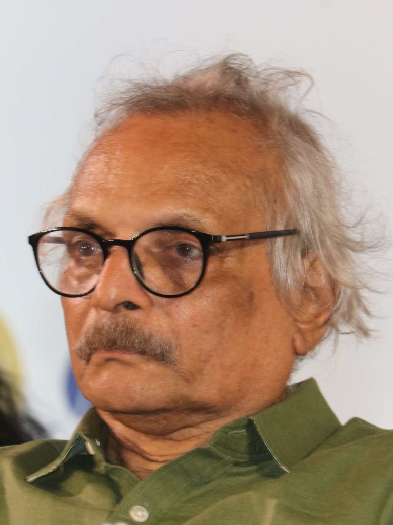M. Mukundan with white hair and a moustache, in round glasses and a green shirt