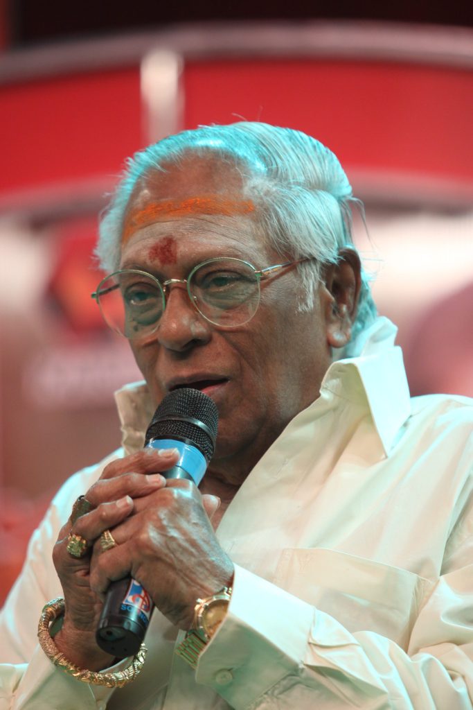 M. S. Viswanathan in a white shirt singing into a microphone
