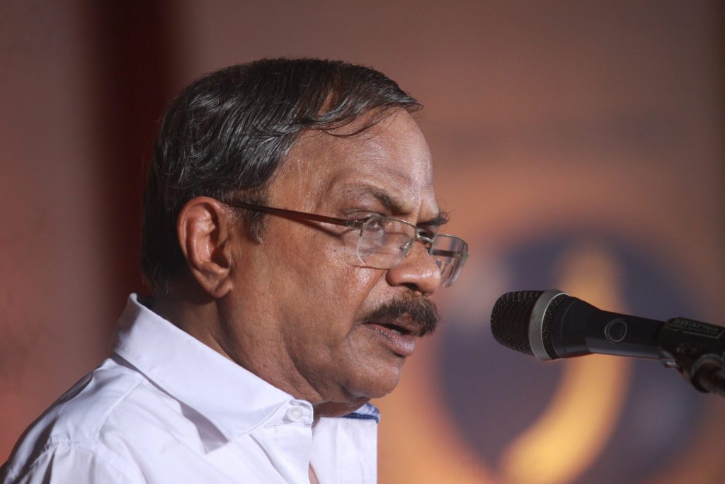 M. T. Vasudevan Nair in glasses and a white shirt speaking at a microphone