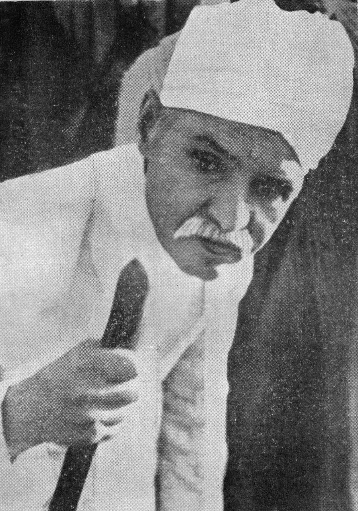 Old photograph of Madan Mohan Malaviya in a white turban and shawl, holding a staff
