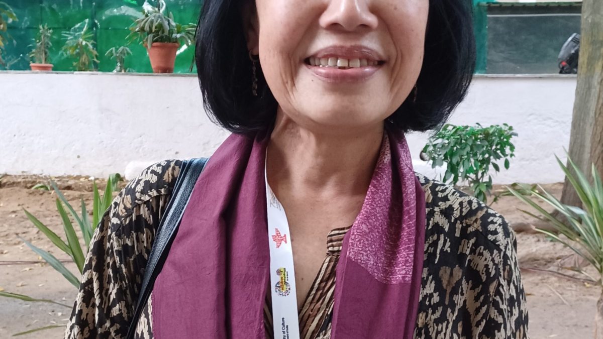 Mamang Dai standing outdoors with a maroon scarf, smiling