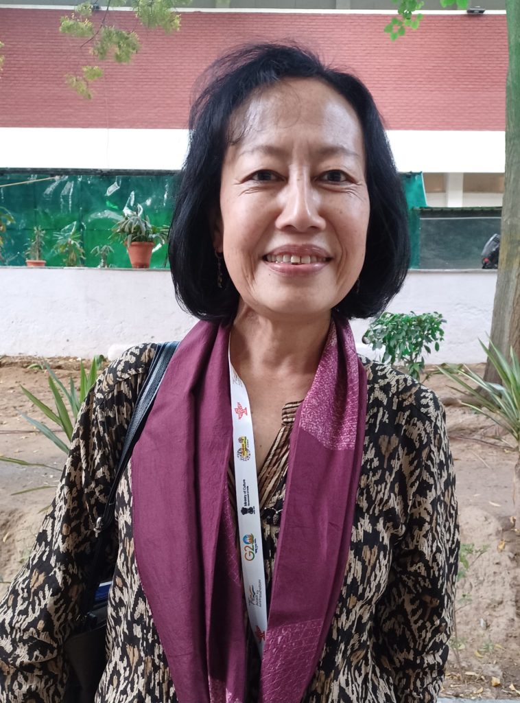 Mamang Dai standing outdoors with a maroon scarf, smiling