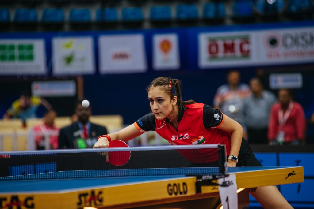 Manika Batra in a red and black shirt leaning over the table to play a shot