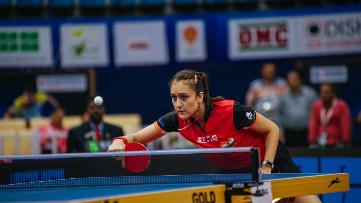 Manika Batra in a red and black shirt leaning over the table to play a shot