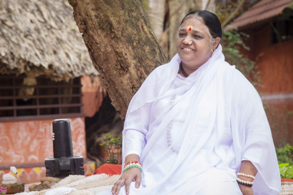Mata Amritanandamayi in white, seated and smiling, with a thatched building behind her