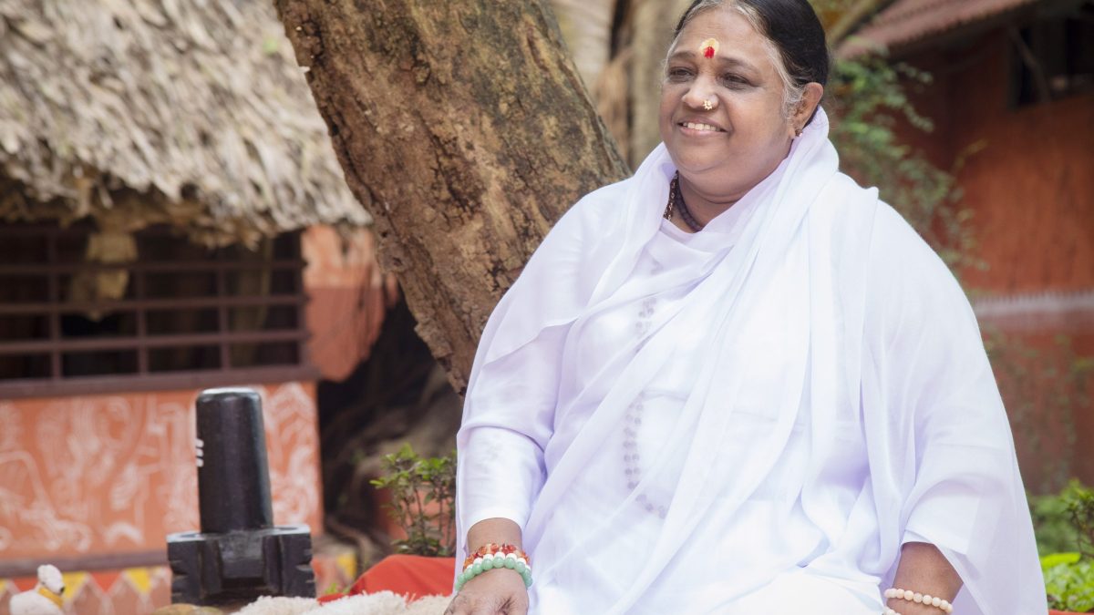 Mata Amritanandamayi in white, seated and smiling, with a thatched building behind her