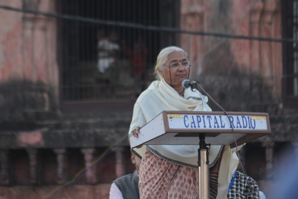 Medha Patkar in a white shawl speaking at a lectern microphone