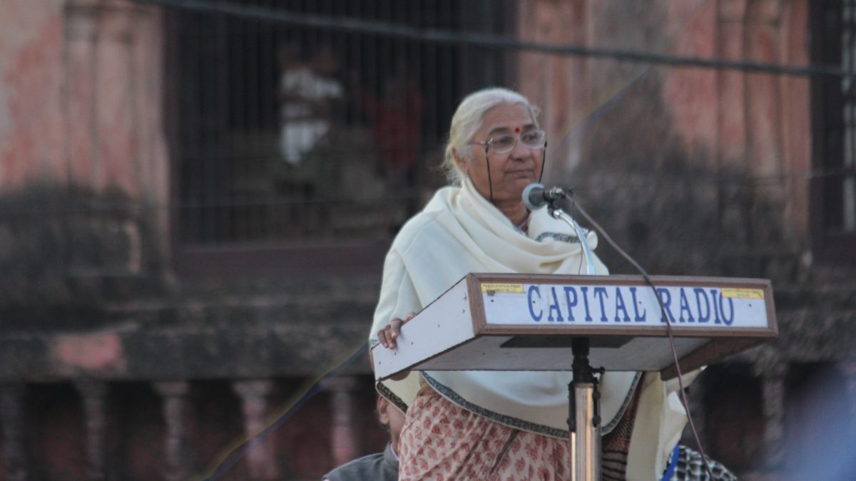 Medha Patkar in a white shawl speaking at a lectern microphone