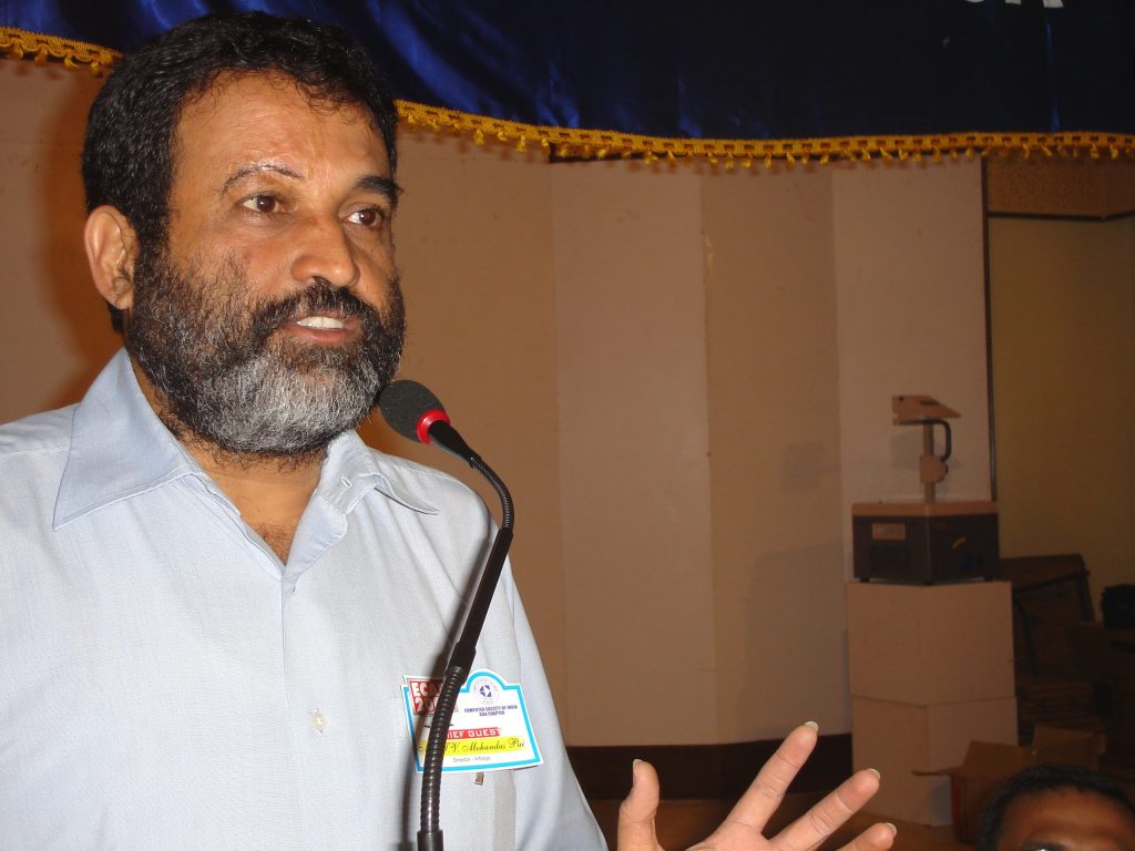 T. V. Mohandas Pai with a beard in a white shirt gesturing as he speaks