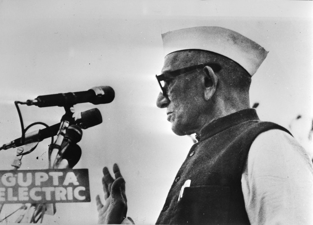 Black and white photo of Morarji Desai in a Gandhi cap and glasses speaking at microphones