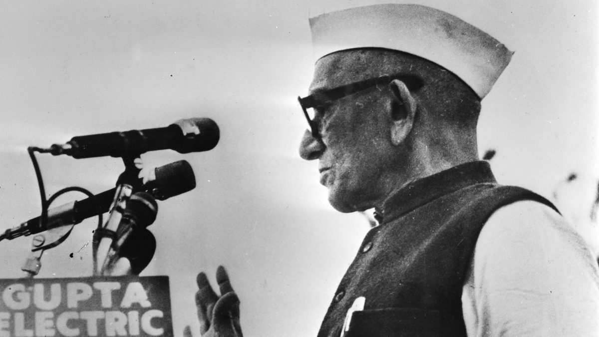 Black and white photo of Morarji Desai in a Gandhi cap and glasses speaking at microphones