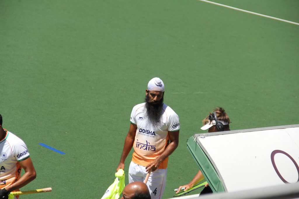 An Indian hockey player in a white kit walking on a green artificial pitch