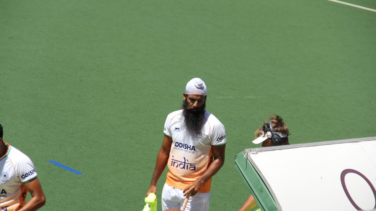 An Indian hockey player in a white kit walking on a green artificial pitch