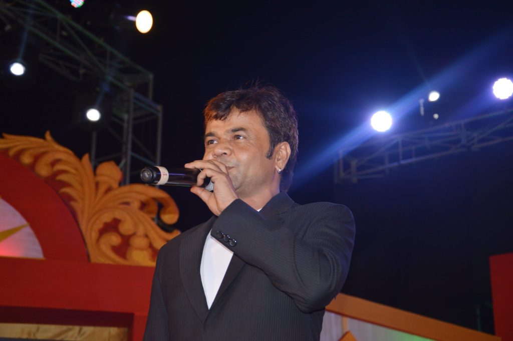Actor Rajpal Yadav in a dark suit speaking into a microphone on stage