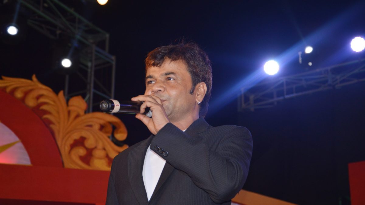 Actor Rajpal Yadav in a dark suit speaking into a microphone on stage