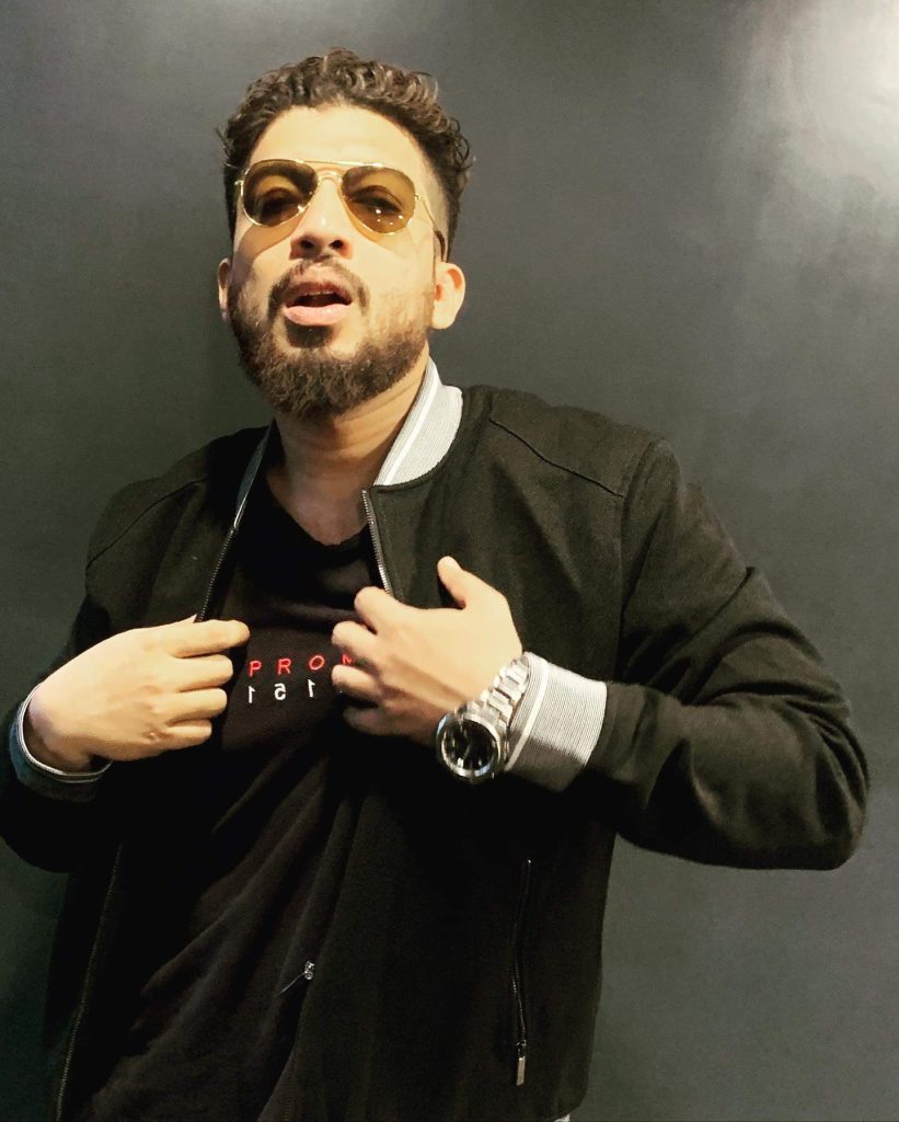 Naezy in a black jacket and tinted glasses, gesturing with both hands