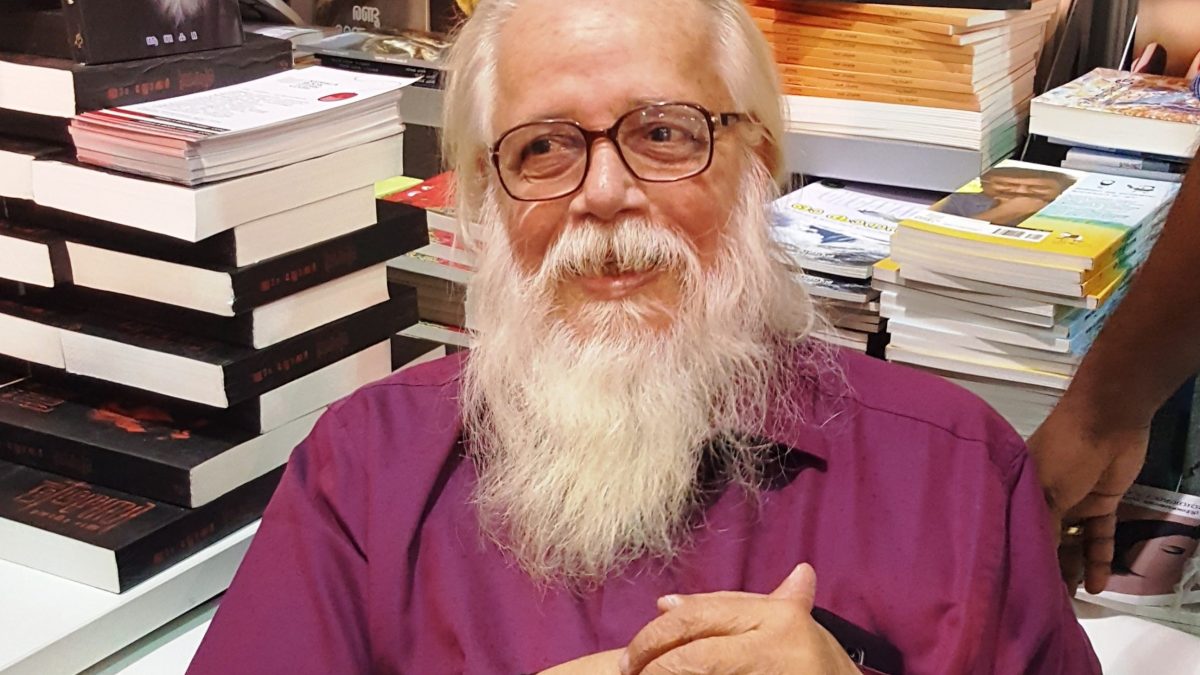 Nambi Narayanan with a white beard, in a purple shirt, seated among books