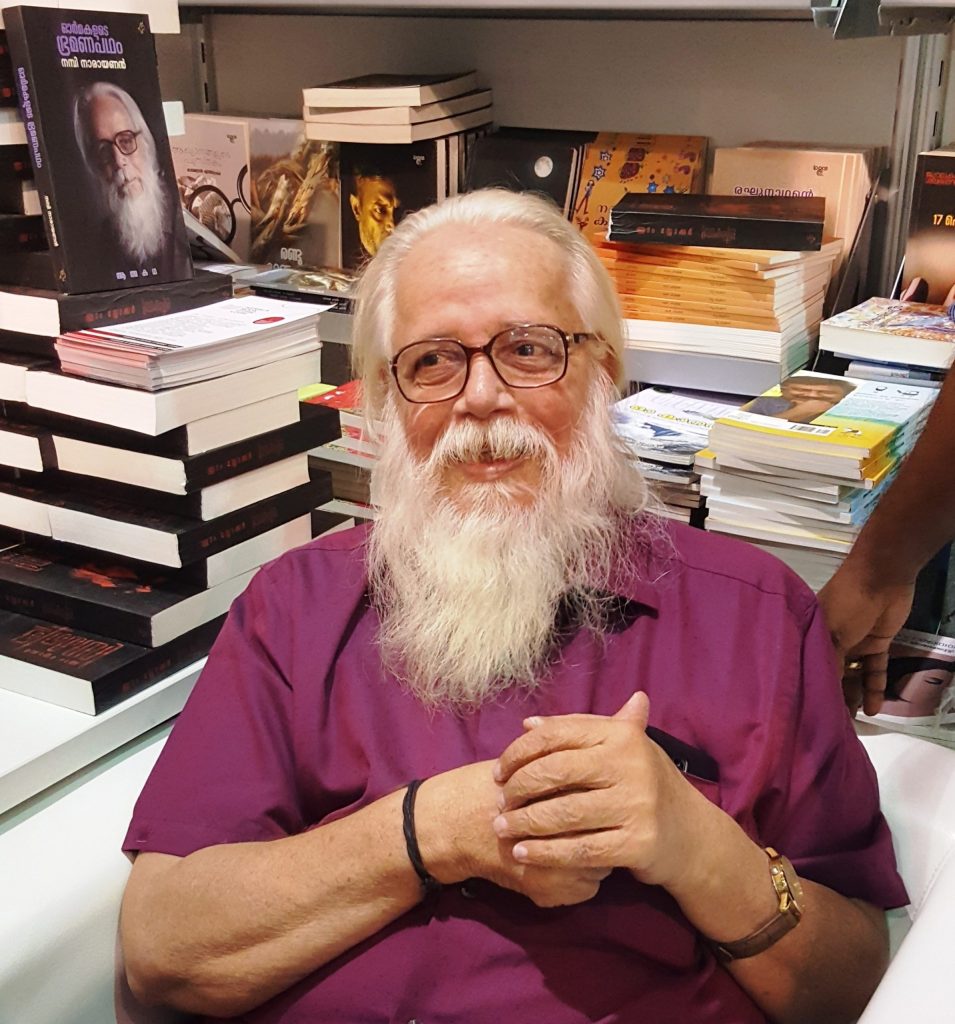 Nambi Narayanan with a white beard, in a purple shirt, seated among books