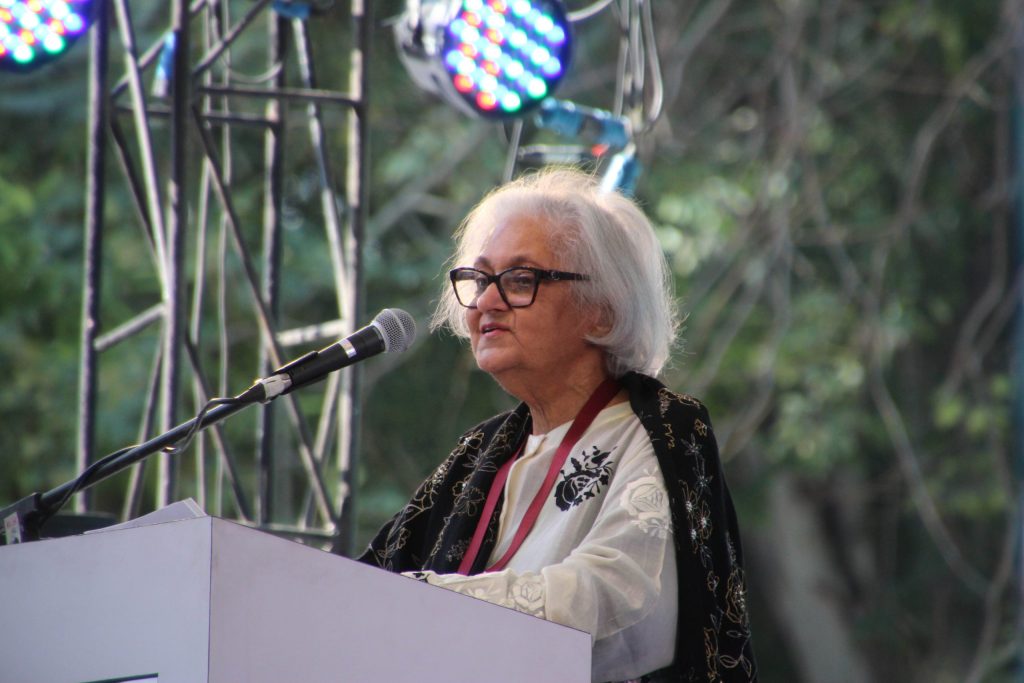 Namita Gokhale with white hair speaking at a lectern microphone