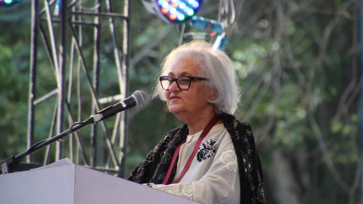 Namita Gokhale with white hair speaking at a lectern microphone