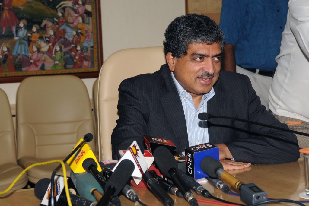 Nandan Nilekani smiling at a desk with press microphones in front of him