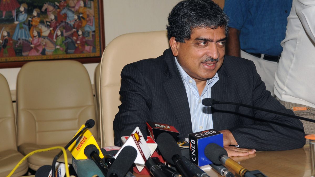 Nandan Nilekani smiling at a desk with press microphones in front of him
