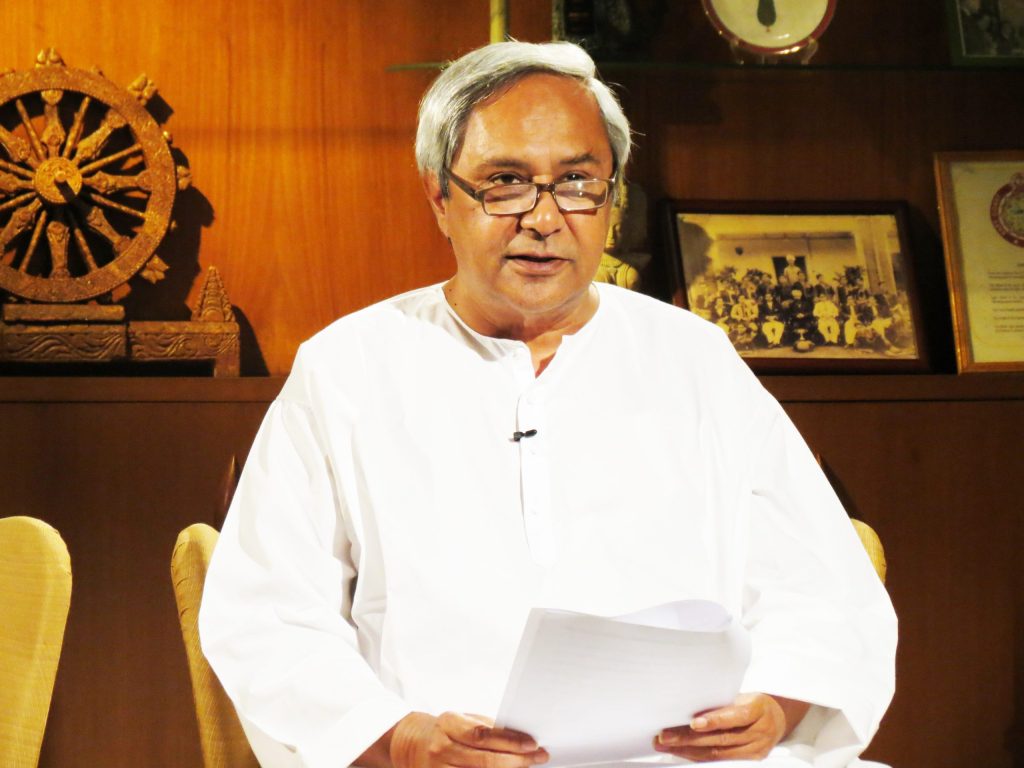 Naveen Patnaik in a white kurta and glasses seated at a desk holding papers