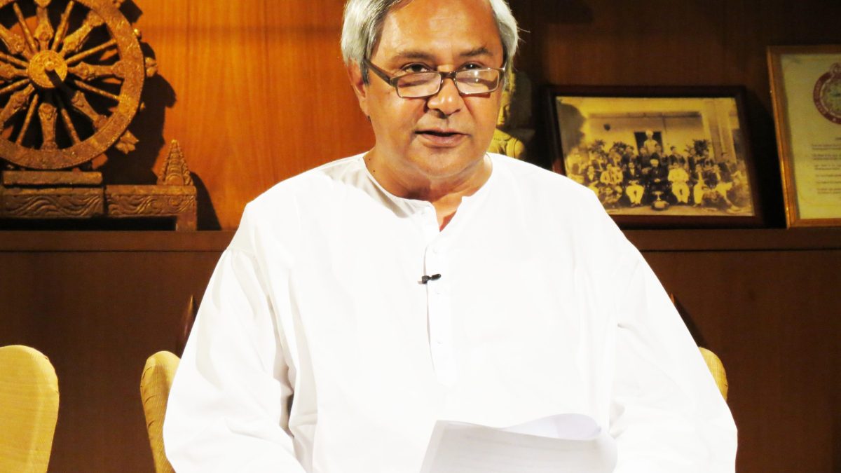 Naveen Patnaik in a white kurta and glasses seated at a desk holding papers