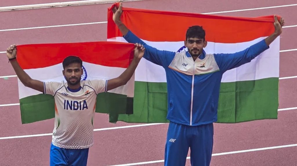 Two Indian athletes in India team kit holding the Indian flag behind them on a running track