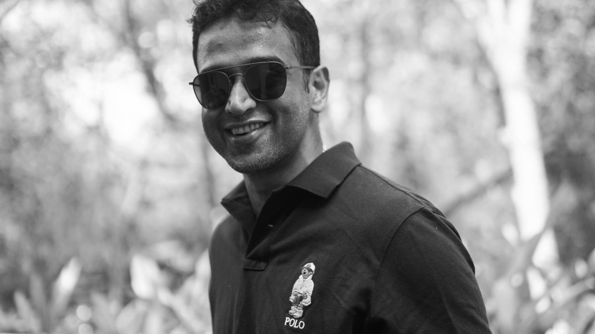 Black and white photo of Nithin Kamath in sunglasses and a polo shirt, sitting outdoors