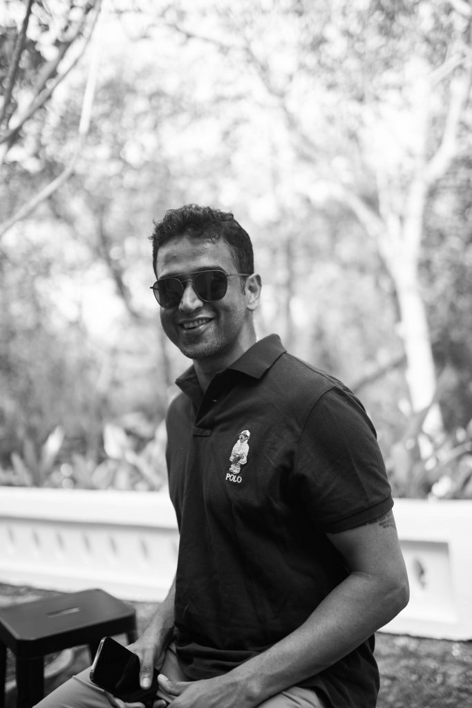 Black and white photo of Nithin Kamath in sunglasses and a polo shirt, sitting outdoors