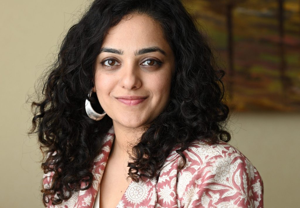 Nithya Menen with curly hair and a silver earring smiling at the camera