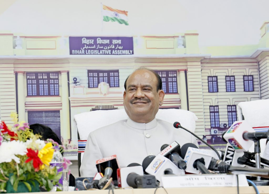 Om Birla in a white kurta and waistcoat smiling as he speaks to the press