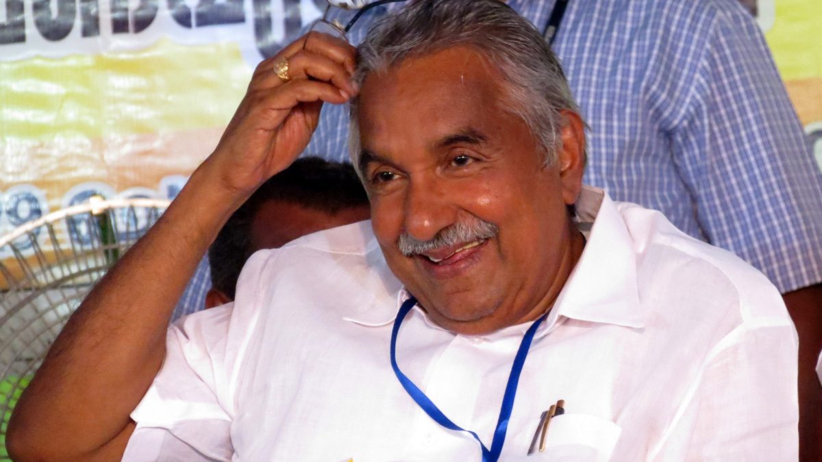 Oommen Chandy with tousled grey hair in a white shirt, smiling and touching his head
