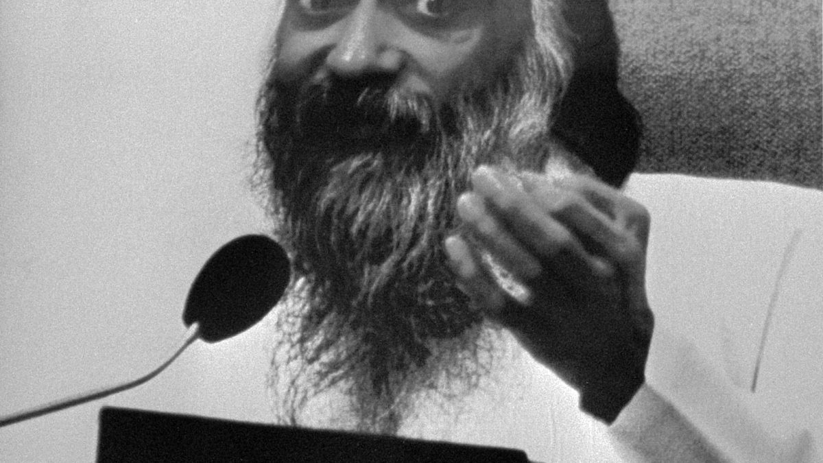 A black-and-white poster portrait of the bearded Bhagwan Shree Rajneesh speaking at a microphone