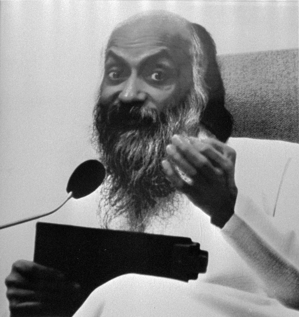 A black-and-white poster portrait of the bearded Bhagwan Shree Rajneesh speaking at a microphone