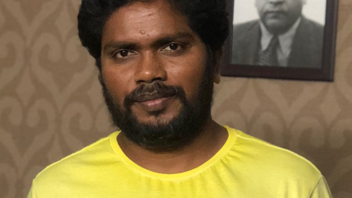 Pa. Ranjith in a yellow T-shirt, with a portrait of B. R. Ambedkar on the wall behind him