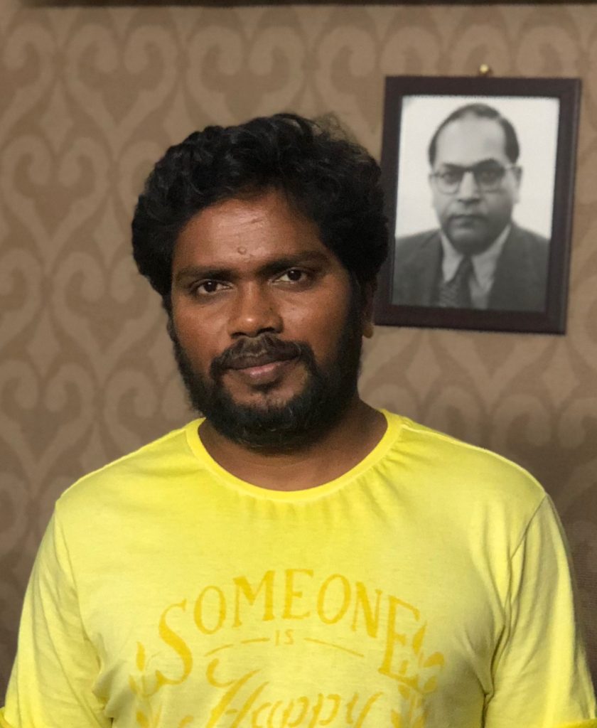 Pa. Ranjith in a yellow T-shirt, with a portrait of B. R. Ambedkar on the wall behind him