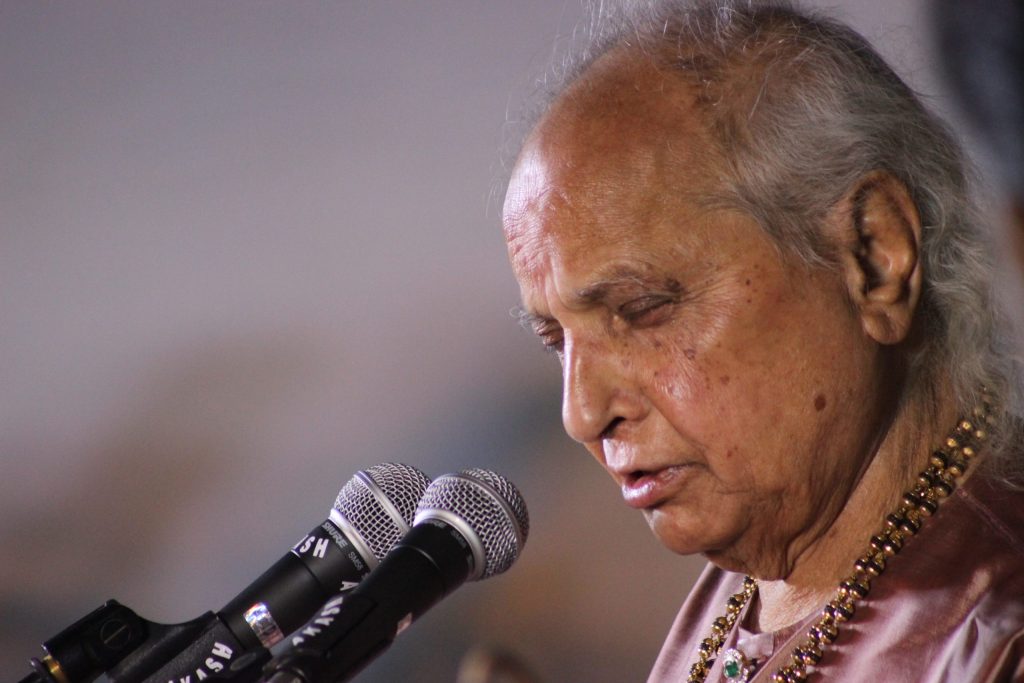 Pandit Jasraj with white hair, eyes closed, singing at microphones