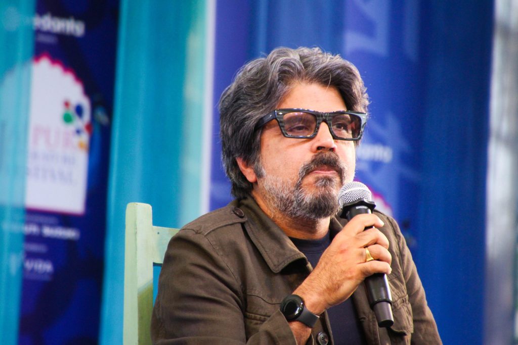 Pankaj Mishra with dark hair and a beard, in a brown jacket, holding a microphone