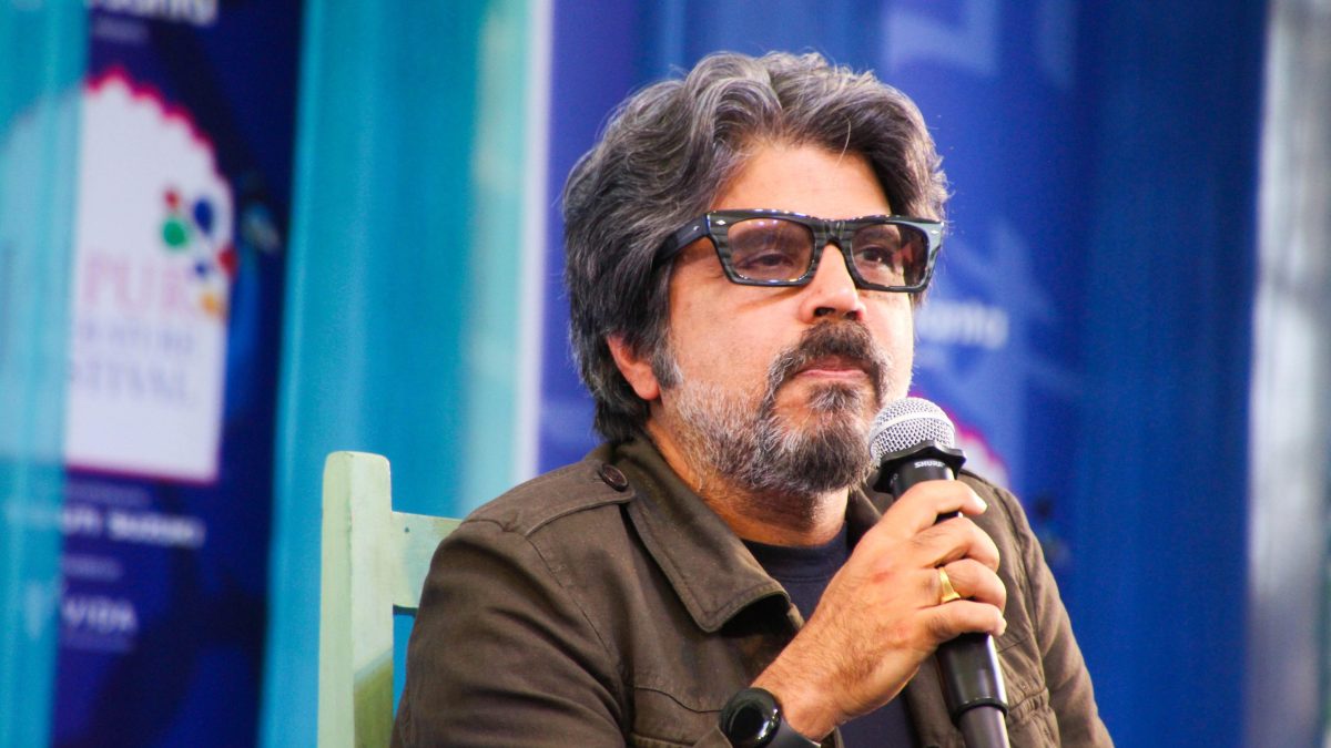 Pankaj Mishra with dark hair and a beard, in a brown jacket, holding a microphone