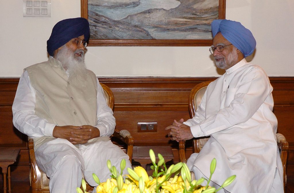 Parkash Singh Badal with a white beard and turban seated in conversation with Prime Minister Manmohan Singh