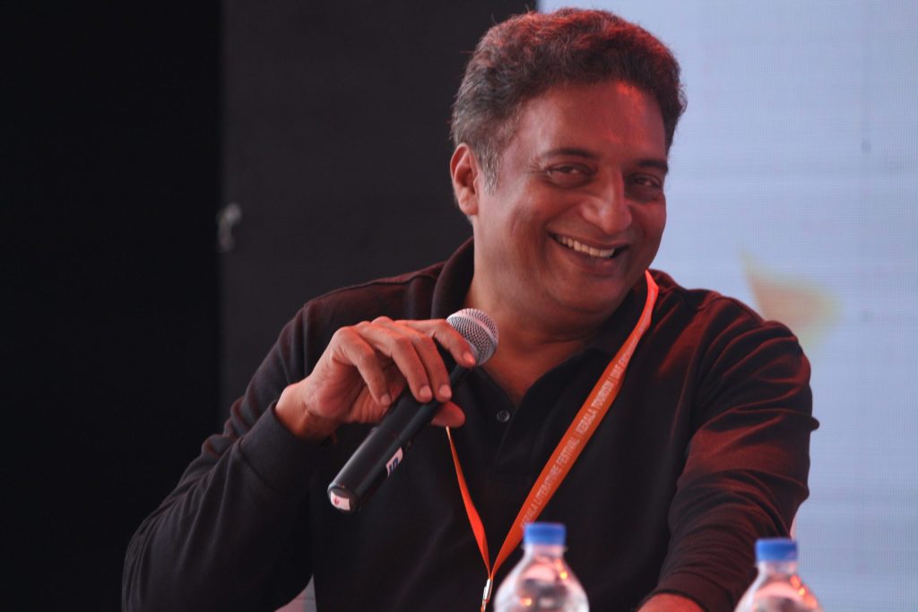 Prakash Raj laughing with a microphone in his hand, wearing a dark shirt and an orange lanyard