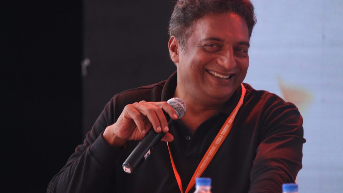 Prakash Raj laughing with a microphone in his hand, wearing a dark shirt and an orange lanyard