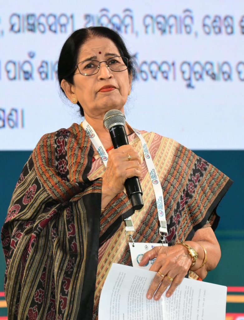 Pratibha Ray in a patterned sari speaking into a microphone on a stage